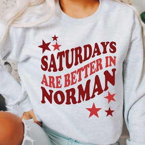 May include: A light gray sweatshirt with the text "Saturdays are better in Norman" in red and maroon lettering. Red stars are scattered around the text. The sweatshirt is a casual style.