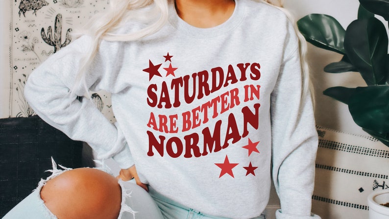 May include: A light gray sweatshirt with the text "Saturdays are better in Norman" in red and maroon lettering. Red stars are scattered around the text. The sweatshirt is a casual style.