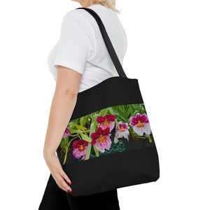 Puede incluir: Una bolsa de tela negra con un diseño floral con orquídeas rosas y blancas. Las orquídeas están en flor y tienen hojas verdes.