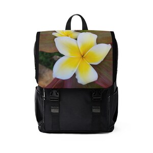 Puede incluir: Mochila negra con un diseño floral con dos flores de plumeria blancas y amarillas sobre un fondo rojo y verde.