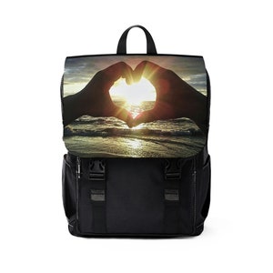 Puede incluir: Mochila negra con una escena de puesta de sol en la solapa delantera. La escena presenta dos manos formando un corazón alrededor del sol poniente sobre el océano.