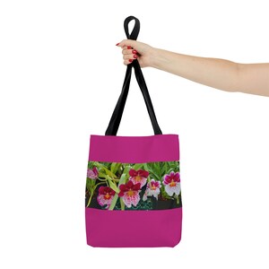 Puede incluir: Una bolsa de mano rosa con correas negras presenta una impresión floral colorida de orquídeas rosas, blancas y verdes.