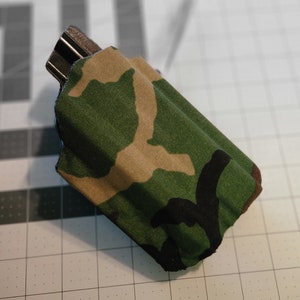 May include: A camouflage fabric pouch with a black zipper. The pouch is folded and lying on a gray grid surface.