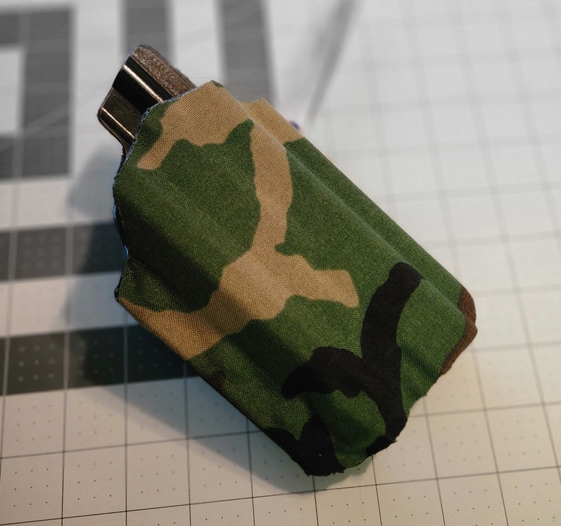 May include: A camouflage fabric pouch with a black zipper. The pouch is folded and lying on a gray grid surface.