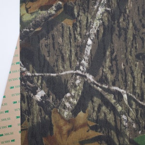 May include: Camouflage fabric with a brown, green, and tan pattern. The fabric has a textured surface and is designed to blend in with the surrounding environment.