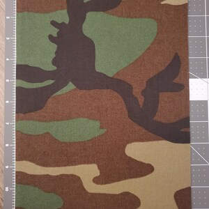 May include: A close-up of a camouflage fabric with brown, green, and tan colors. The fabric has a woven texture and is suitable for crafting projects.