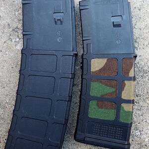 May include: Two black 5.56 x 45 magazines. One magazine has a camouflage pattern covering the front and sides. The text "5.56 x 45" is printed on the top of both magazines.