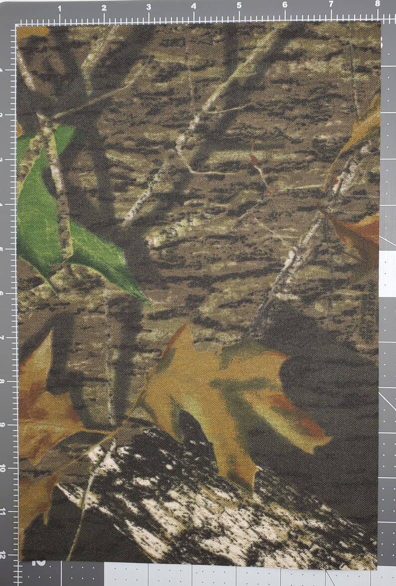 May include: Camouflage fabric with a brown, green, and tan pattern. The fabric has a woven texture and a subtle sheen.