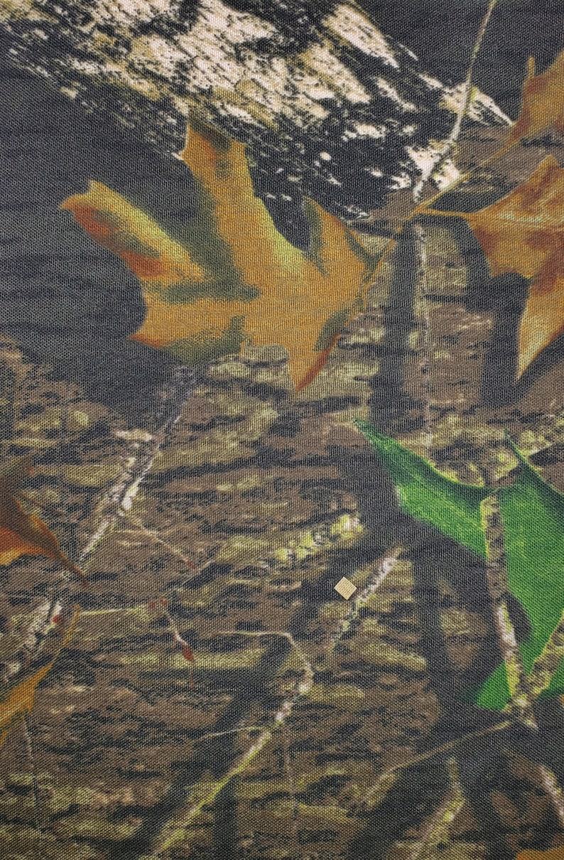 May include: Camouflage fabric with a brown, green, and white pattern. The fabric features a textured weave and a realistic depiction of leaves and branches.