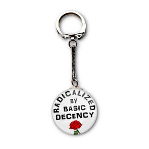 May include: A white keychain with the text "RADICALIZED BY BASIC DECENCY" in black, with a red rose graphic. The keychain has a silver-colored ring and chain.