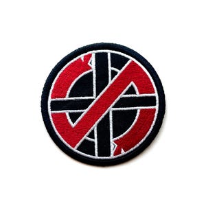 May include: Embroidered patch featuring a black and red design with a white border. The design includes a red diagonal line crossing a black circle with a cross and arrows. The patch has a navy blue border and is circular in shape.