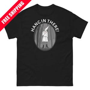 May include: Black t-shirt with a graphic of a figure in a white robe and hood, with a red heart, hanging in a forest scene. The text "HANG IN THERE!" is above the image. A red banner with "FREE SHIPPING" is in the upper left corner.