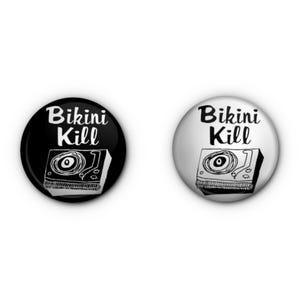 May include: Two round buttons with the text "Bikini Kill" above a drawing of a record player. One button is black with white text and image, the other is white with black text and image.