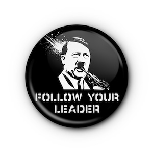 May include: A black button with a white graphic of a man holding a gun and the words "FOLLOW YOUR LEADER" in white block letters. The graphic has a splatter effect above the man's head.