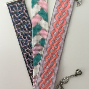 Könnte beinhalten: Drei Armbänder mit Perlen sind abgebildet. Eines hat ein dunkelblaues geometrisches Muster, ein anderes rosa, weiße und türkisfarbene Streifen, und das dritte ein korallenfarbenes und lavendelfarbenes Design. Jedes Armband hat einen silbernen Verschluss.