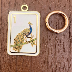 K&ouml;nnte beinhalten: Ein rechteckiger wei&szlig;er Emaille-Anh&auml;nger mit goldfarbenem Rand und einer Pfauen-Illustration auf einem gr&uuml;nen Bambuszweig mit der Zahl 1 in der Ecke, begleitet von einem runden ros&eacute;goldfarbenen Metallring zur Befestigung.