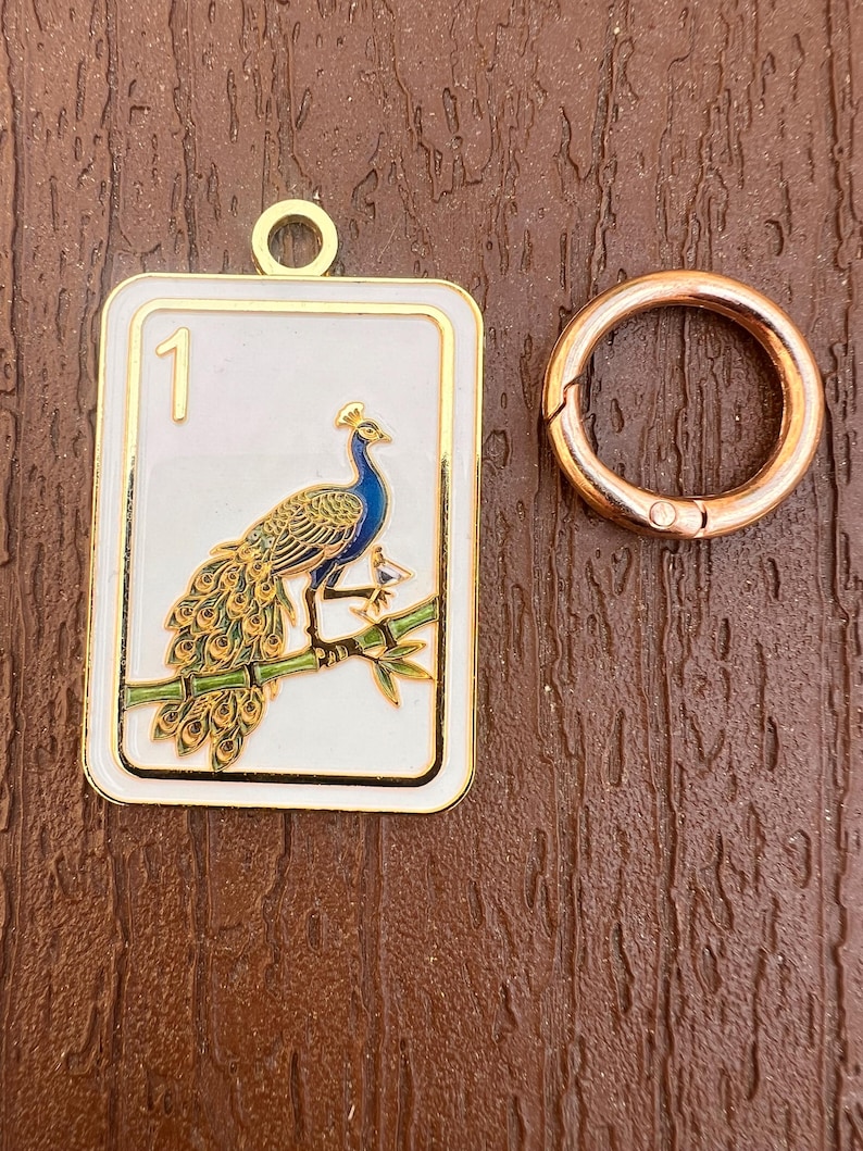 K&ouml;nnte beinhalten: Ein rechteckiger wei&szlig;er Emaille-Anh&auml;nger mit goldfarbenem Rand und einer Pfauen-Illustration auf einem gr&uuml;nen Bambuszweig mit der Zahl 1 in der Ecke, begleitet von einem runden ros&eacute;goldfarbenen Metallring zur Befestigung.