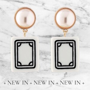 May include: A pair of white and black rectangular earrings with a gold-colored top and a pearl-like bead. The earrings have a black border and a design in the center. The text "NEW IN" is at the bottom.