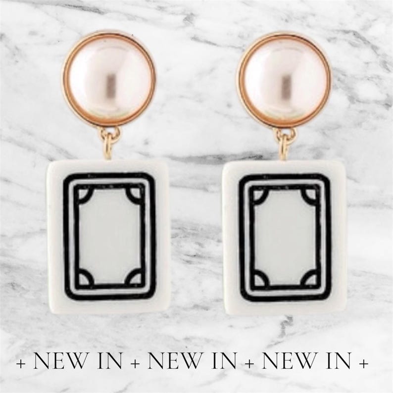 May include: A pair of white and black rectangular earrings with a gold-colored top and a pearl-like bead. The earrings have a black border and a design in the center. The text "NEW IN" is at the bottom.