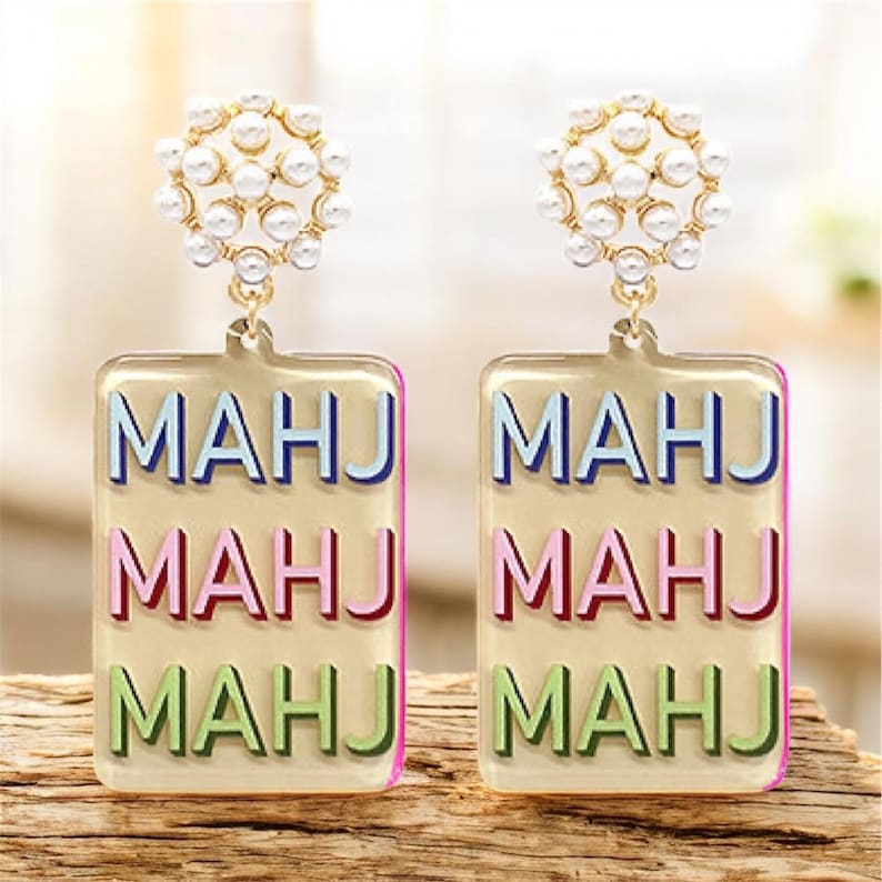 May include: A pair of gold-toned earrings. Each earring features a cluster of small, white faux pearls at the top, connected to a rectangular pendant. The pendant has the word "MAHJ" in blue, pink, and green.