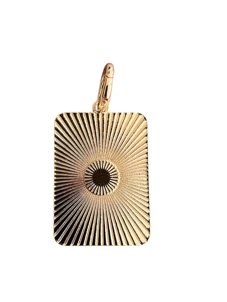 May include: Gold-colored rectangular pendant with a sunburst design. The pendant has a circular center and radiating lines. A small gold clasp is attached at the top. The pendant is likely made of metal and is suitable for jewelry.