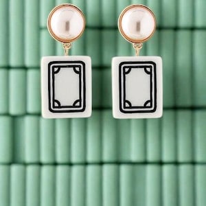 May include: A pair of gold-toned earrings with a pearl stud and a white rectangular pendant featuring a black border design. The earrings are displayed against a light green background.