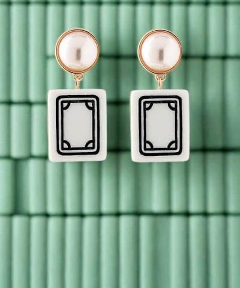 May include: A pair of gold-toned earrings with a pearl stud and a white rectangular pendant featuring a black border design. The earrings are displayed against a light green background.