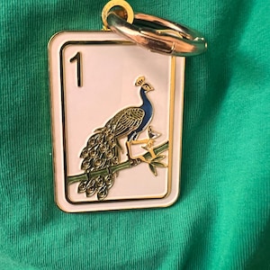 K&ouml;nnte beinhalten: Ein rechteckiger Metallanh&auml;nger mit wei&szlig;em Emaillehintergrund und goldenem Rand. Das Motiv zeigt einen Pfau auf einem gr&uuml;nen Bambuszweig mit der Zahl 1 in der oberen linken Ecke. Ein goldfarbener, runder Verschluss ist an der Oberseite befestigt.