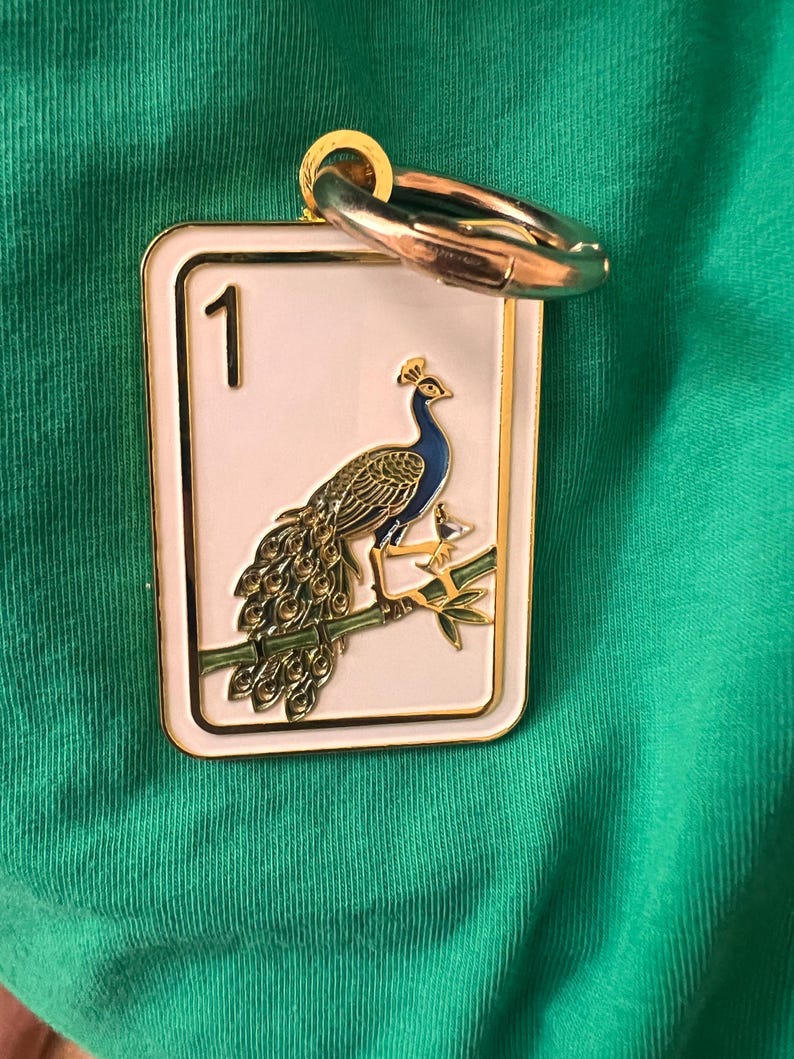 K&ouml;nnte beinhalten: Ein rechteckiger Metallanh&auml;nger mit wei&szlig;em Emaillehintergrund und goldenem Rand. Das Motiv zeigt einen Pfau auf einem gr&uuml;nen Bambuszweig mit der Zahl 1 in der oberen linken Ecke. Ein goldfarbener, runder Verschluss ist an der Oberseite befestigt.