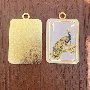 K&ouml;nnte beinhalten: Zwei rechteckige Metallanh&auml;nger mit goldfarbenen &Ouml;sen an der Oberseite. Ein Anh&auml;nger hat eine texturierte goldene Oberfl&auml;che. Der andere zeigt einen goldfarbenen Pfau auf einem gr&uuml;nen Bambuszweig vor wei&szlig;em Emaille-Hintergrund und die Zahl 1.