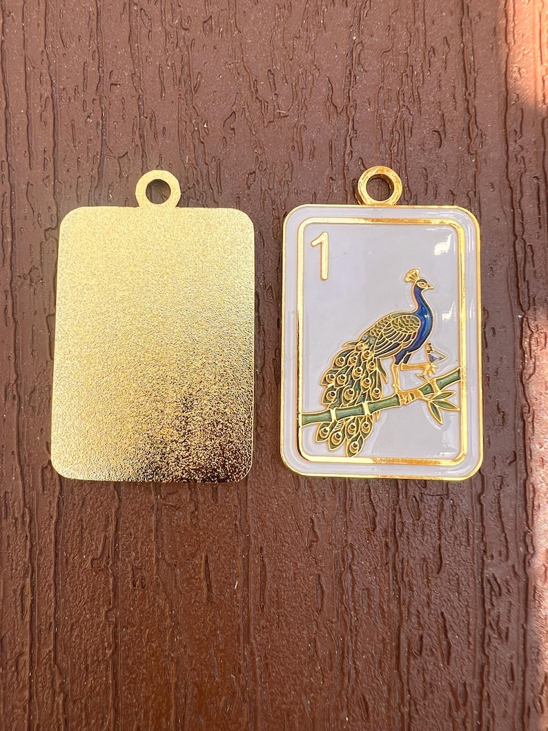 K&ouml;nnte beinhalten: Zwei rechteckige Metallanh&auml;nger mit goldfarbenen &Ouml;sen an der Oberseite. Ein Anh&auml;nger hat eine texturierte goldene Oberfl&auml;che. Der andere zeigt einen goldfarbenen Pfau auf einem gr&uuml;nen Bambuszweig vor wei&szlig;em Emaille-Hintergrund und die Zahl 1.