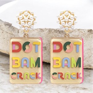 K&ouml;nnte beinhalten: Ein Paar bunte Statement-Ohrringe. Jeder Ohrring hat einen rechteckigen, transparenten Acryl-Anh&auml;nger mit den Worten "DOT BAM CRACK" in bunten, geschichteten Buchstaben. Die Oberseite jedes Ohrrings hat eine Ansammlung kleiner, wei&szlig;er Kunstperlen.