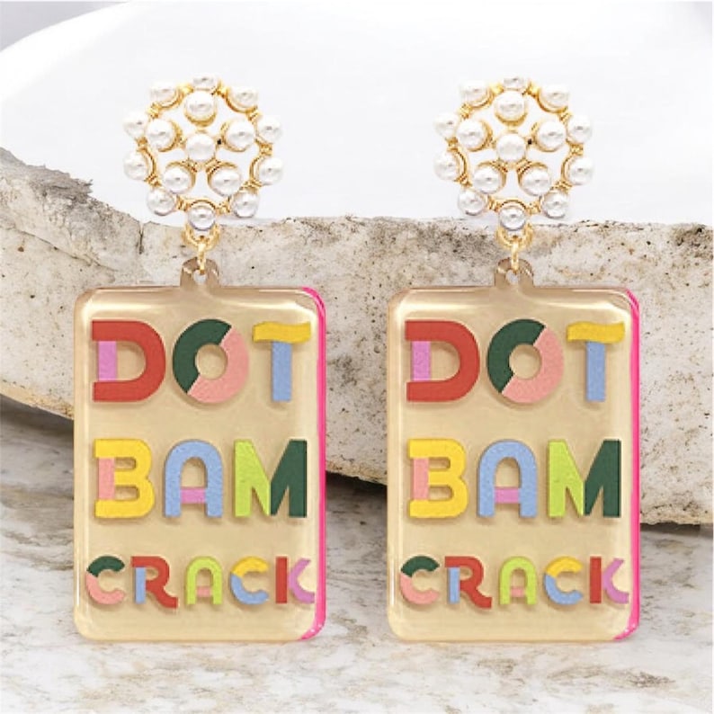 K&ouml;nnte beinhalten: Ein Paar bunte Statement-Ohrringe. Jeder Ohrring hat einen rechteckigen, transparenten Acryl-Anh&auml;nger mit den Worten "DOT BAM CRACK" in bunten, geschichteten Buchstaben. Die Oberseite jedes Ohrrings hat eine Ansammlung kleiner, wei&szlig;er Kunstperlen.