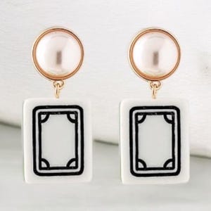 May include: A pair of earrings featuring a gold-colored circular top with a pearl-like center, connected to a white rectangular charm with a black border. The charm has a rectangular design in the center.