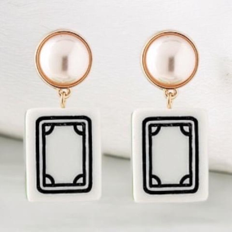 May include: A pair of earrings featuring a gold-colored circular top with a pearl-like center, connected to a white rectangular charm with a black border. The charm has a rectangular design in the center.