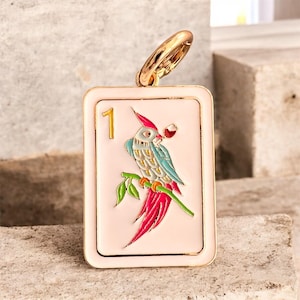 May include: A rectangular pendant with a gold-colored ring at the top. The pendant has a light pink background with a gold border. It features a colorful parrot perched on a branch, holding a glass of wine. The number "1" is in the upper left corner.