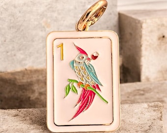 May include: A rectangular pendant with a gold-colored ring at the top. The pendant has a light pink background with a gold border. It features a colorful parrot perched on a branch, holding a glass of wine. The number "1" is in the upper left corner.