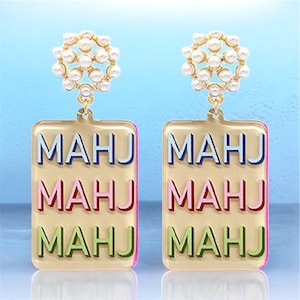 May include: A pair of gold-toned earrings. Each earring features a cluster of small, white pearls at the top. Below, a rectangular pendant displays the word "MAHJ" in blue, pink, and green. The earrings have a pink border.