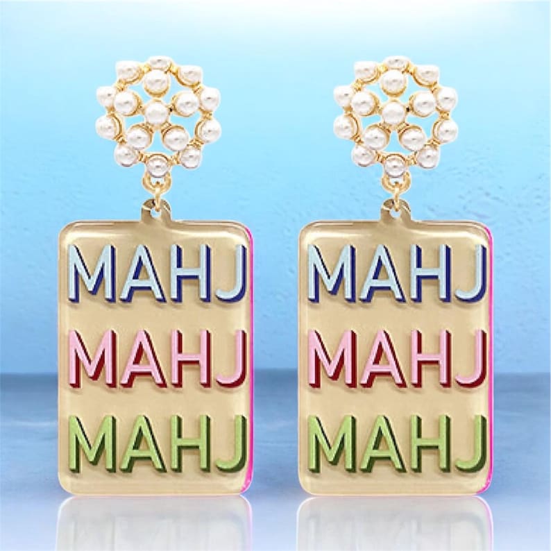 May include: A pair of gold-toned earrings. Each earring features a cluster of small, white pearls at the top. Below, a rectangular pendant displays the word "MAHJ" in blue, pink, and green. The earrings have a pink border.