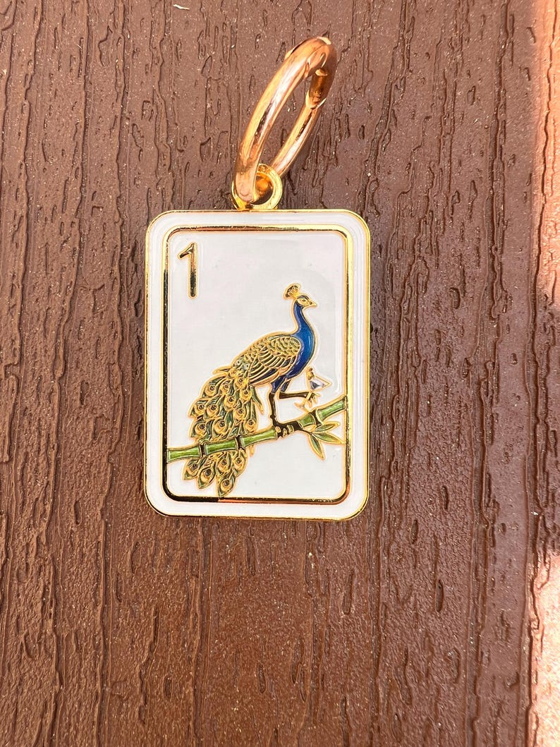K&ouml;nnte beinhalten: Ein rechteckiger Anh&auml;nger aus goldfarbenem Metall mit wei&szlig;em Emaille-Hintergrund und goldenem Rand. Das Motiv zeigt einen Pfau auf einem gr&uuml;nen Bambuszweig, mit der Zahl 1 in der oberen linken Ecke. Ein goldener Ring ist an der Oberseite befestigt.