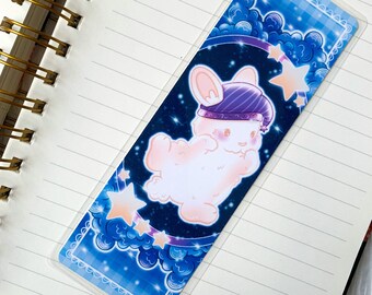 May include: A white bunny wearing a purple and white hat with a star on top, floating in a blue and white starry sky with clouds. The bunny is surrounded by stars and clouds. The bookmark is made of clear plastic.