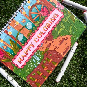 May include: A spiral-bound coloring book with the words "HAPPY COLORING" on the cover. The cover features a colorful garden scene with flowers, a wooden wheel, and string lights. Several white markers with colored tips surround the book on a bed of green grass.