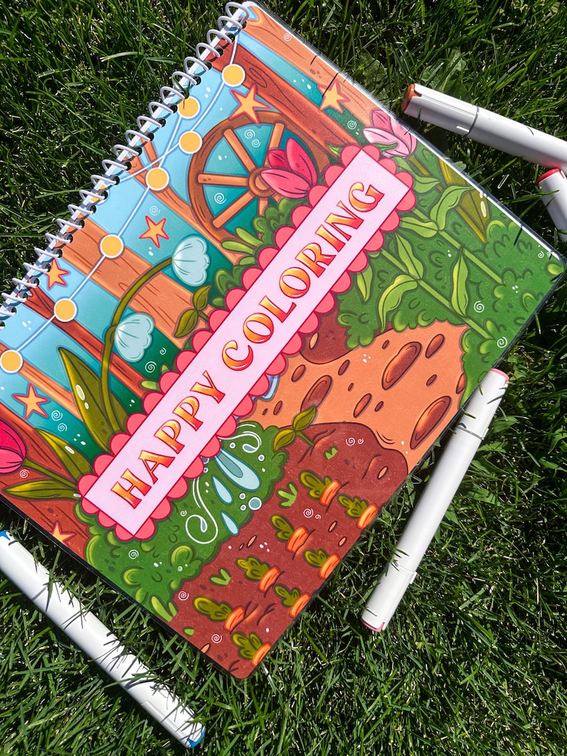 May include: A spiral-bound coloring book with the words "HAPPY COLORING" on the cover. The cover features a colorful garden scene with flowers, a wooden wheel, and string lights. Several white markers with colored tips surround the book on a bed of green grass.