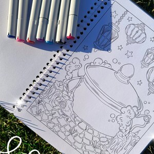 May include: A spiral-bound coloring book with a black and white illustration of a cauldron, lanterns, and stars. Seven white markers with colored tips are arranged on the open book. The text "MAGIC CAULDRON" is visible.