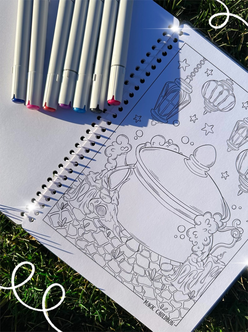 May include: A spiral-bound coloring book with a black and white illustration of a cauldron, lanterns, and stars. Seven white markers with colored tips are arranged on the open book. The text "MAGIC CAULDRON" is visible.