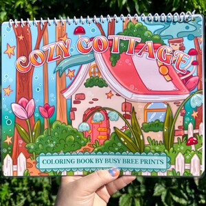 May include: A spiral-bound coloring book titled "Cozy Cottage" with a whimsical illustration of a cottage, flowers, and trees. The cover features the text "COLORING BOOK BY BUSY BREE PRINTS". The color palette includes pink, blue, green, and orange.