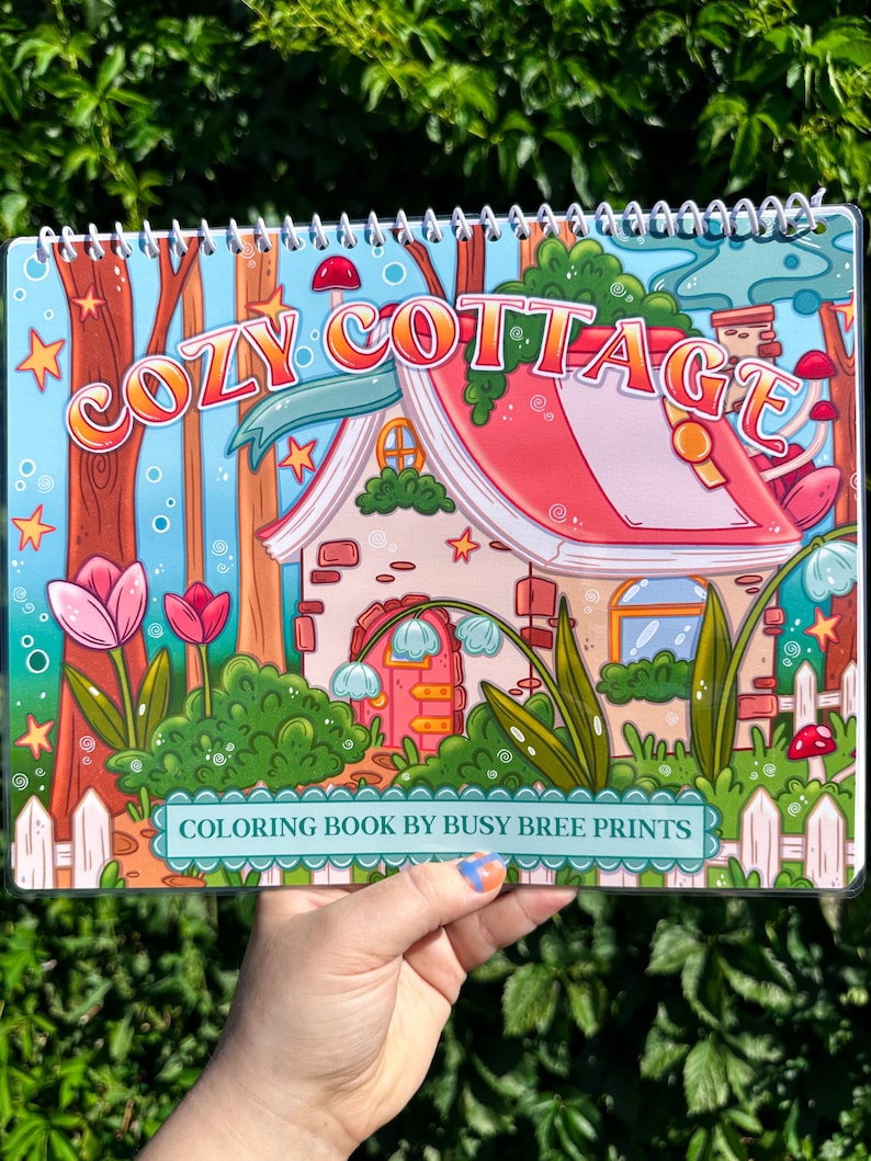May include: A spiral-bound coloring book titled "Cozy Cottage" with a whimsical illustration of a cottage, flowers, and trees. The cover features the text "COLORING BOOK BY BUSY BREE PRINTS". The color palette includes pink, blue, green, and orange.