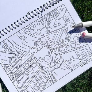 May include: A black and white line art coloring book with a detailed interior scene, including a computer, shelves, and floral elements. Two markers are visible. The book is open on a bed of green grass.