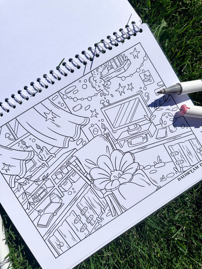 May include: A black and white line art coloring book with a detailed interior scene, including a computer, shelves, and floral elements. Two markers are visible. The book is open on a bed of green grass.