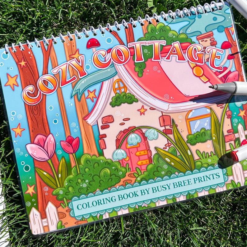 May include: A spiral-bound coloring book with the title "COZY COTTAGE" in orange letters. The cover features a colorful illustration of a cottage, trees, and flowers. The text "COLORING BOOK BY BUSY BREE PRINTS" is at the bottom.